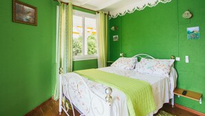 Un dormitorio con paredes verdes, una cama con ropa de cama floral, una ventana con cortinas y una mesita de noche pequeña.