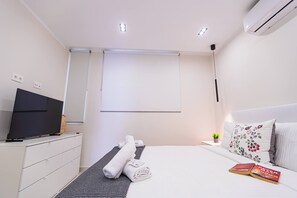 A modern hotel room with a bed, a TV, a bedside table with a lamp, and a window with blinds. - Beachfront apartment in El Duque (Costa Adeje)