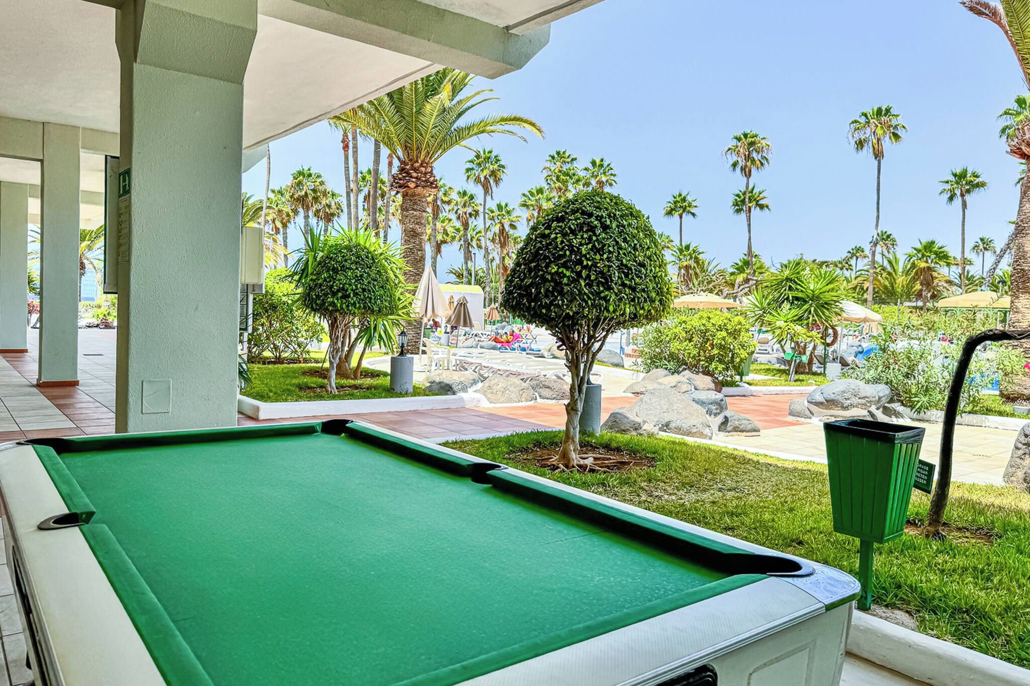 A pool table in a covered area with a view of a landscaped outdoor area including palm trees and a pool.