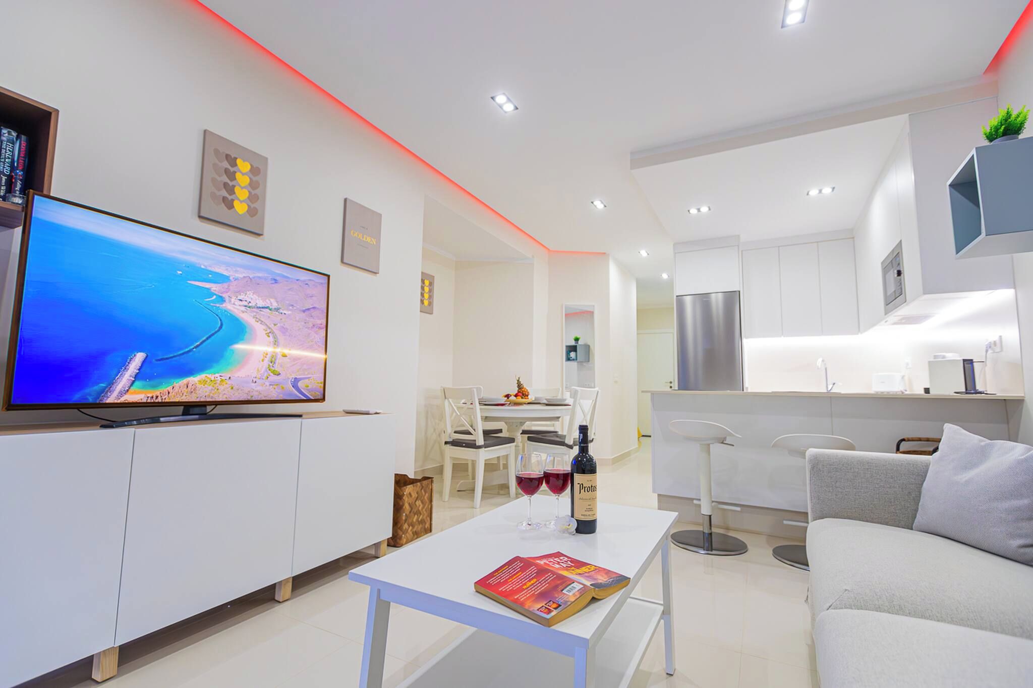 A modern living room with a flat-screen TV, a dining area with a table and chairs, and a kitchen with a refrigerator and bar stools.