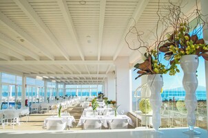 Un comedor amplio con mesas y sillas blancas, ventanas grandes con vistas al mar y un techo con diseño de cuadrícula.