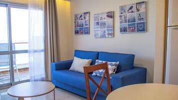 Een woonkamer met een blauwe bank, een houten stoel, een klein rond tafeltje en een groot raam met gordijnen.