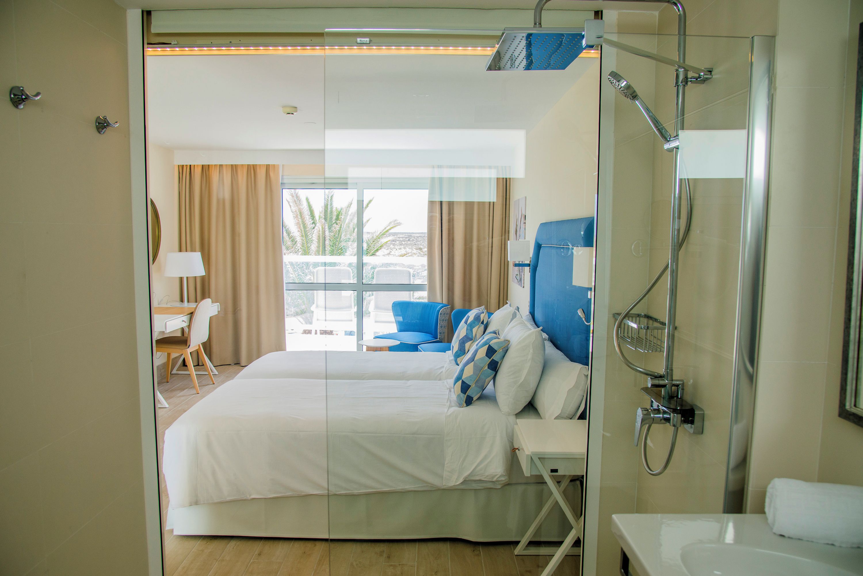 Habitación de hotel con cama, escritorio, silla, baño con ducha y vistas a palmeras.