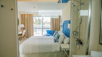 Habitación de hotel con cama, escritorio, silla, baño con ducha y vistas a palmeras.