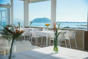 A restaurant with white tables and chairs, set against a backdrop of a clear blue sea and distant mountains. - Hotel Boutique Tao Caleta Mar (La Oliva)