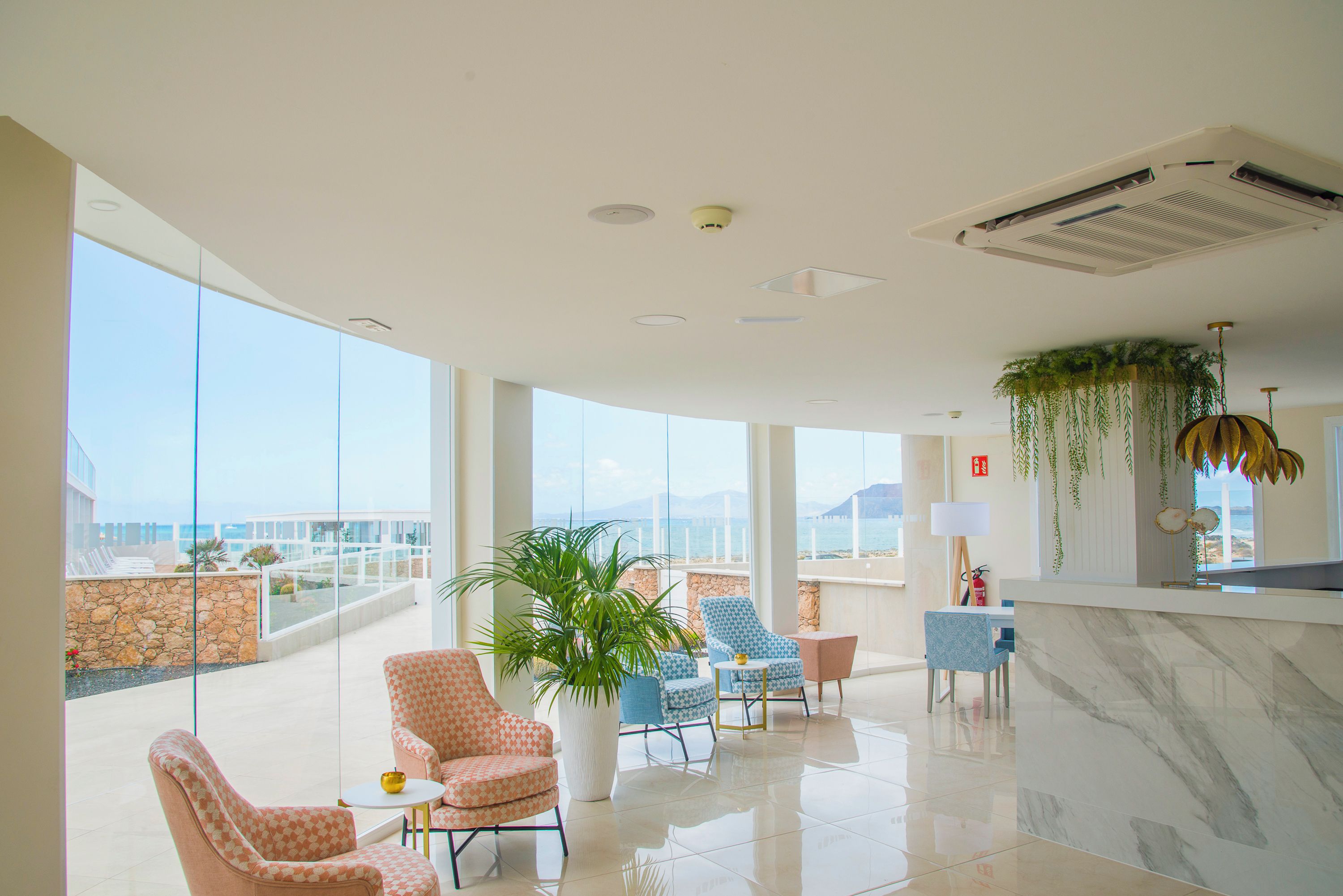 Eine geräumige Lobby mit hoher Decke, großen Fenstern und Meerblick. Es gibt mehrere Sitzbereiche mit gemusterten Polstern und eine große Kühlpflanze.