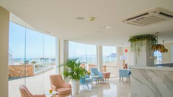 Eine geräumige Lobby mit hoher Decke, großen Fenstern und Meerblick. Es gibt mehrere Sitzbereiche mit gemusterten Polstern und eine große Kühlpflanze.