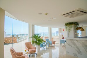 Un vestíbulo amplio con techo alto, ventanas grandes y vista al mar. Varias zonas de estar con tapicería estampada y una gran planta en maceta.