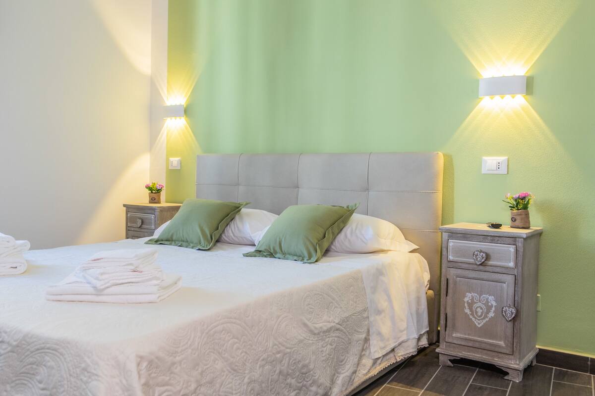 Ein ordentlich bezogenes Bett mit weißer Daunendecke und grünen Kissen, ein Nachttisch mit Pflanze sowie eine an der Wand befestigte Leuchte.