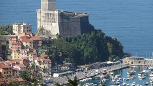 Une ville côtière avec un château imposant perché sur une colline, un port de plaisance rempli de bateaux et des bâtiments aux couleurs vives.