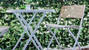 Une table et une chaise en métal blanc se dressent sur fond d’un bâtiment blanc aux volets verts, le tout étant entouré d’une haie verdoyante et dense.