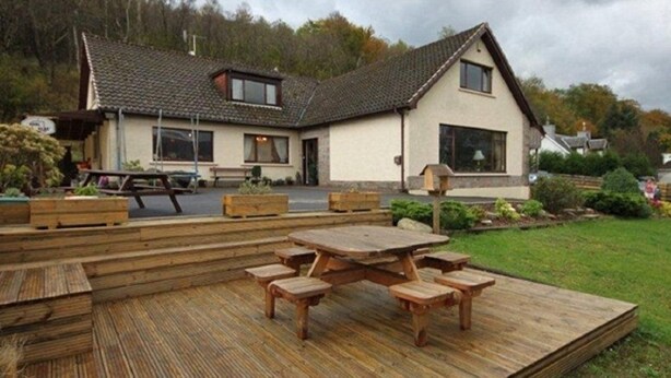 A two-story house with a sloped roof, a front porch with a wooden bench, and a wooden deck with a picnic table. - Stronchreggan View Guest House (Fort William)