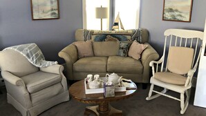 Ein gemütliches Wohnzimmer mit einem beigen Sofa, einem weißen Schaukelstuhl und einem kleinen, runden Tisch mit Teeservice.