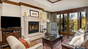 A living room with a fireplace, a television, a patterned armchair, a sofa, and a large window with a view of trees.
