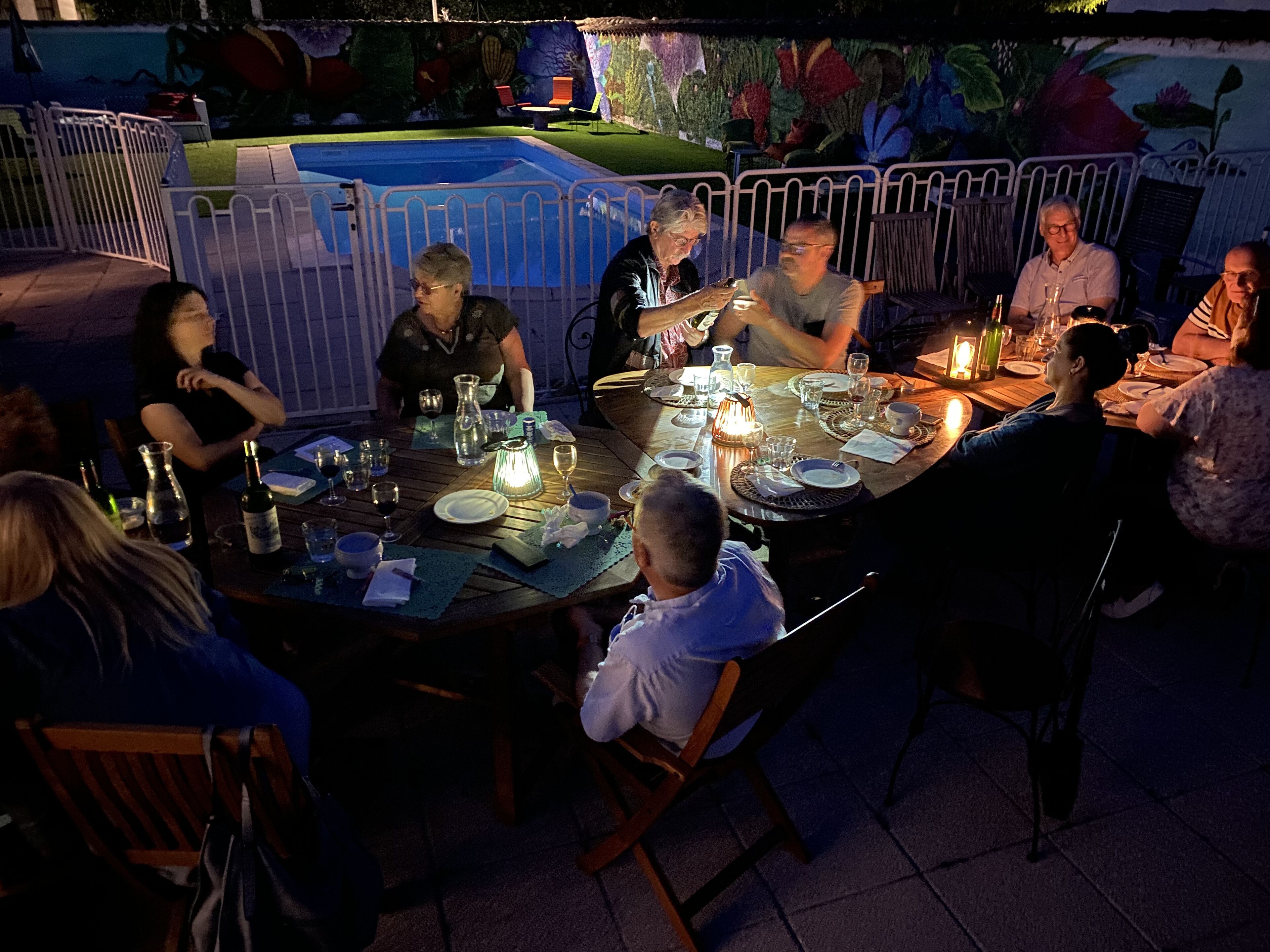 Un groupe de personnes dînant en plein air, de nuit, autour de tables dressées pour le repas, le tout étant entouré d’une clôture décorative et avec une piscine en arrière-plan.