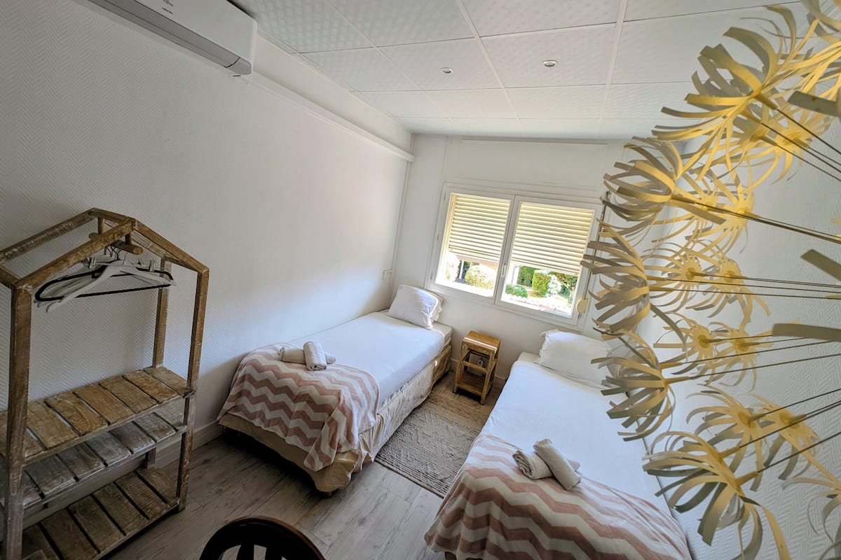 A hotel room with two beds, a wooden bench, a small table, and a window with blinds.