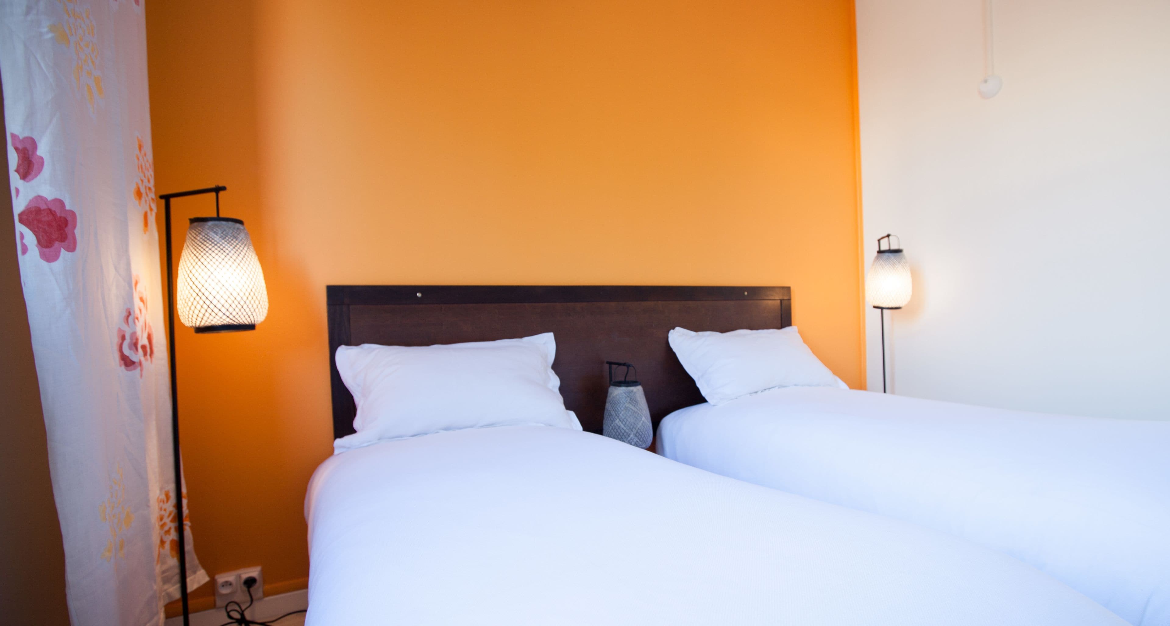 Habitación de hotel con dos camas, cabecero de madera, mesita de noche con lámpara y cortina con motivos florales.