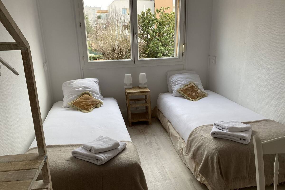 A bedroom with two beds, a wooden nightstand with two lamps, and a window with a view of buildings and trees.