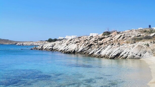 Une plage avec une eau d’un bleu limpide, un rivage rocheux et des bâtiments blancs en arrière-plan.