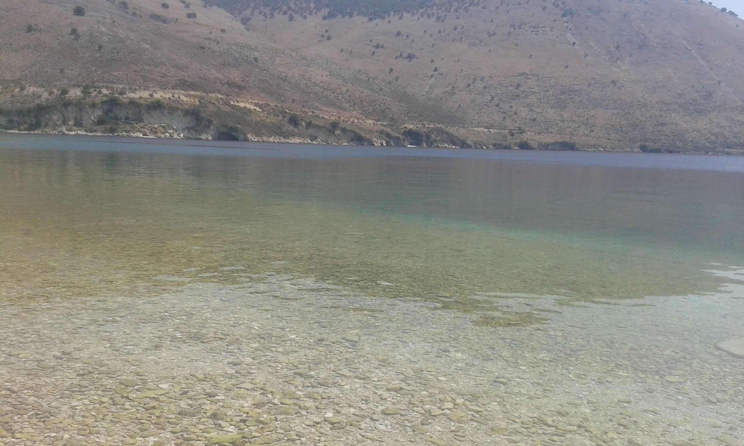 Un lago transparente con piedras visibles en la orilla y un paisaje montañoso al fondo.