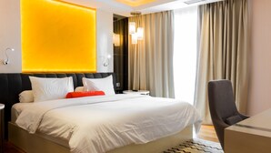 Hotelkamer met een groot bed, een bureau en een stoel. Er is een nachtkastje met een lamp en een spiegel. De kamer heeft een modern design met een geel accentmuur.