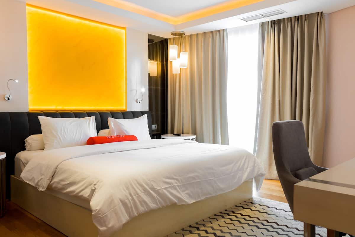 Habitación de hotel con una cama grande, un escritorio y una silla. Hay una mesita de noche con una lámpara y un espejo. La habitación tiene un diseño moderno con una pared de acento amarilla.