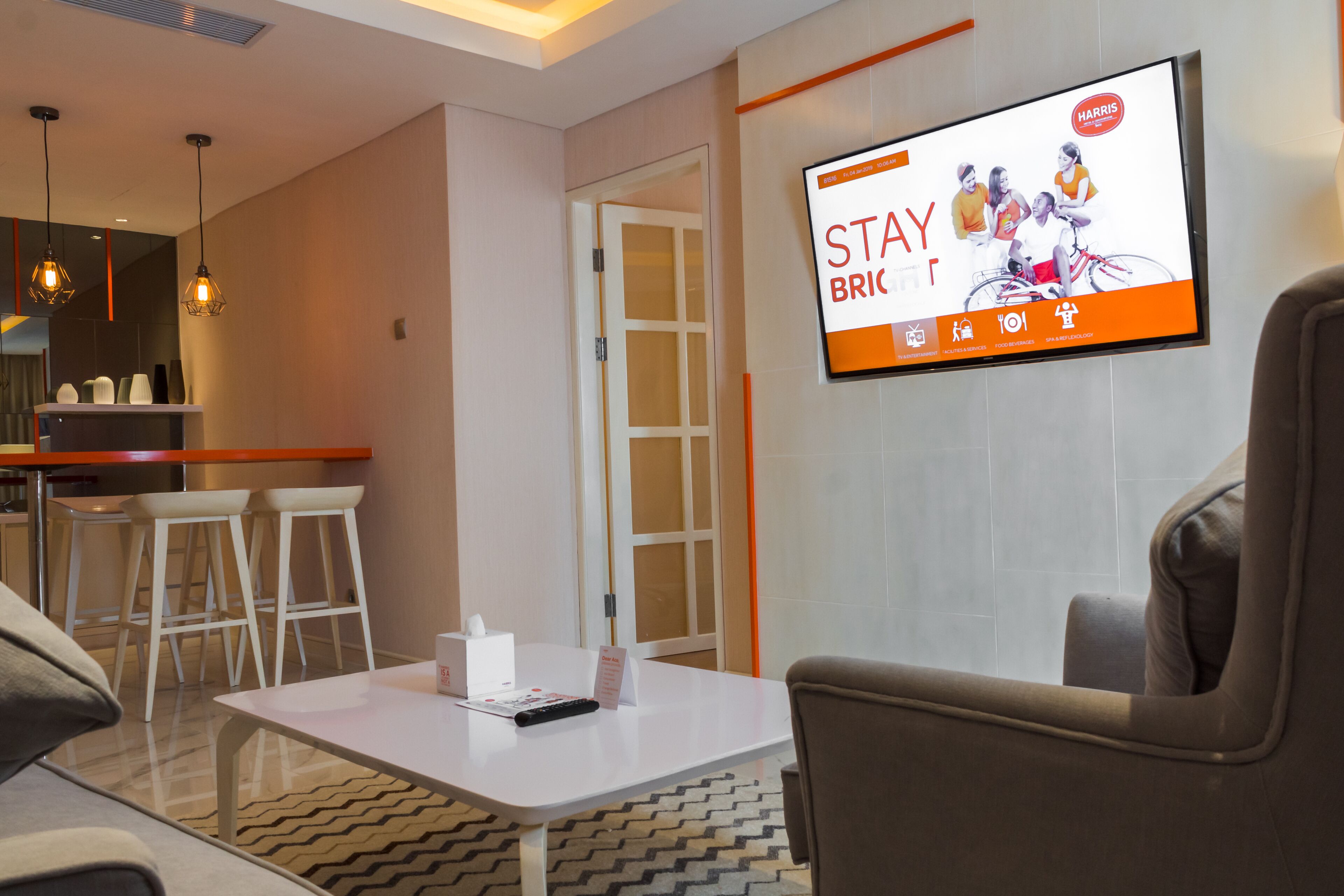 A modern hotel lobby with a flat-screen TV displaying an advertisement, a white reception desk, a bar area, and seating arrangements.