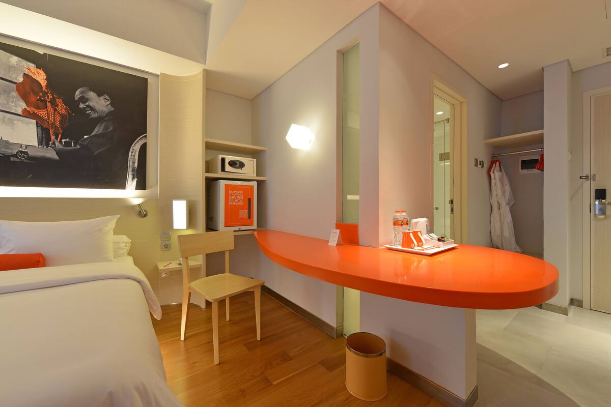 Una habitación de hotel con cama, escritorio, silla, una mesita con bandeja, cesto de basura, perchero y un cuadro en la pared.