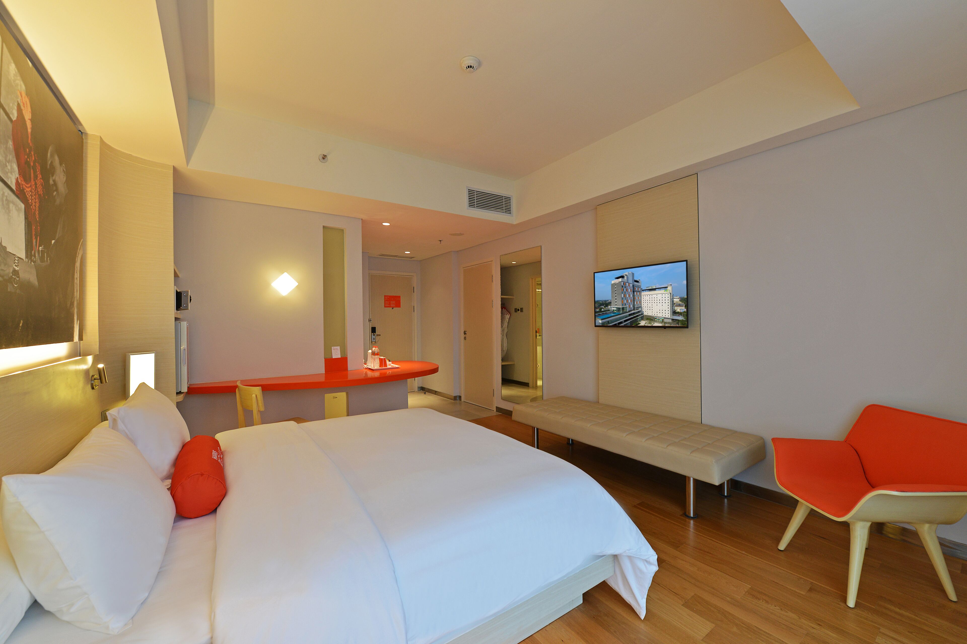 Una habitación de hotel moderna con una cama grande, un banco, un televisor y una silla rojo brillante.