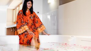 Een persoon in een oranje gewaad, zittend in een badkuip vol rozenblaadjes.