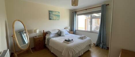 A bedroom with a bed, a bedside table, a mirror, a window with curtains, and a painting on the wall.