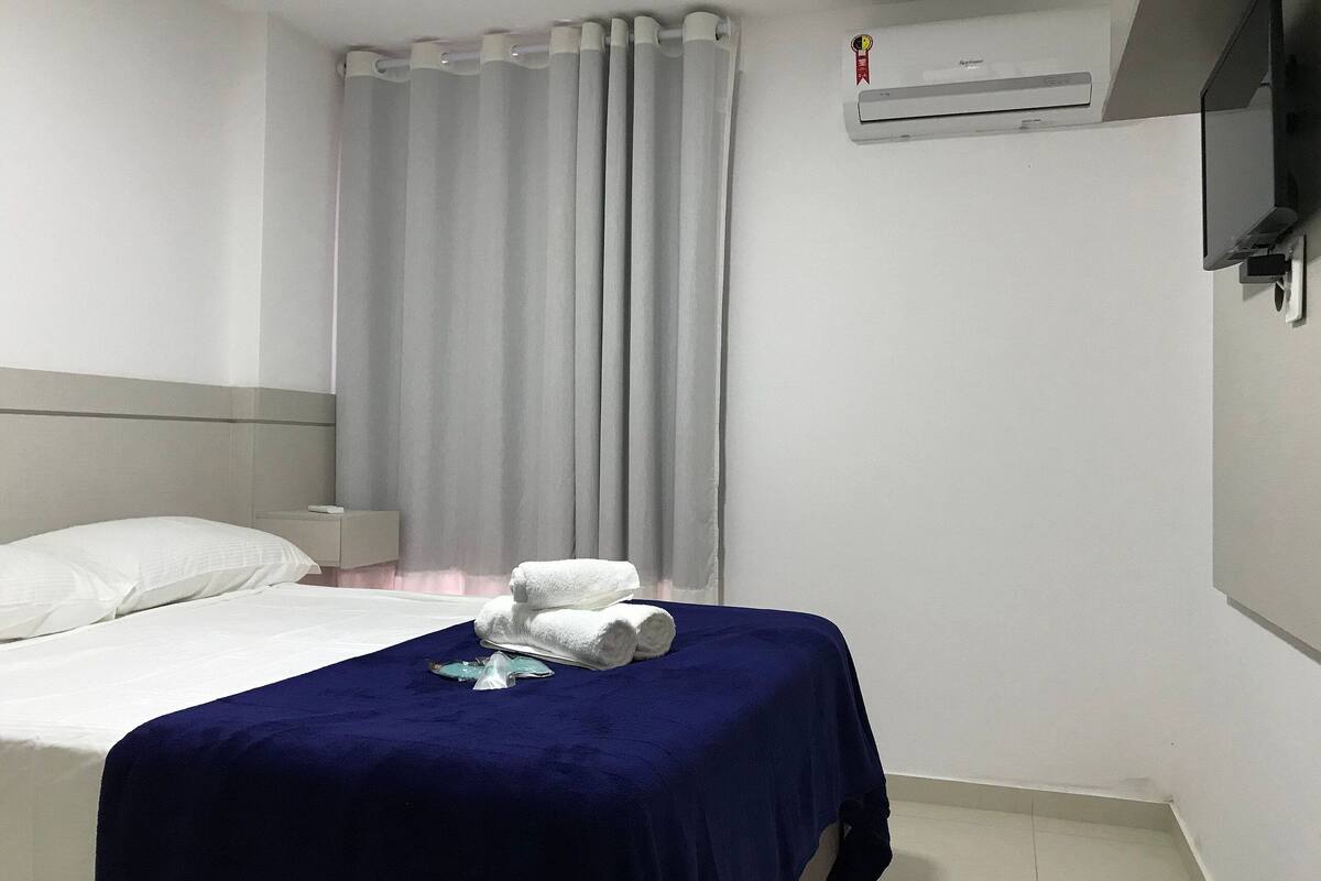 Una habitación de hotel con una cama, una colcha azul, una almohada blanca, una toalla blanca, una cortina gris y un equipo de aire acondicionado.