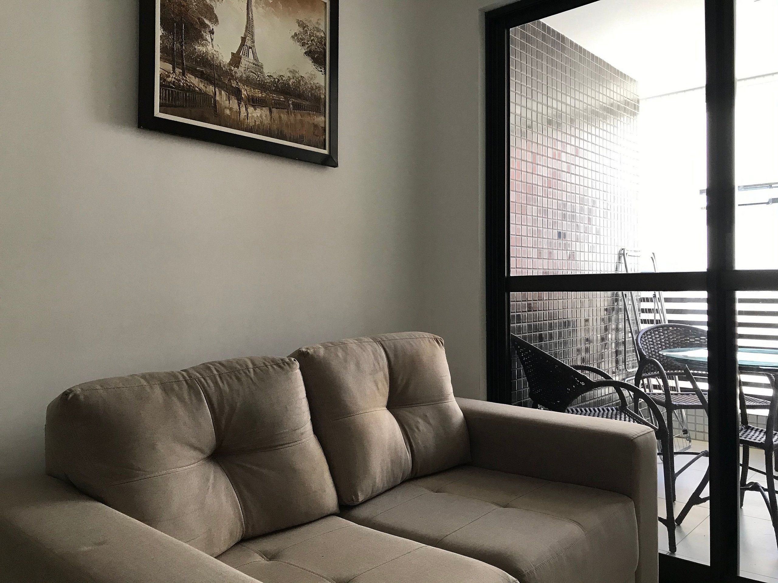 Een beige bank in een kamer met een groot raam, een lijst met een foto van de Eiffeltoren en uitzicht op een gebouw.
