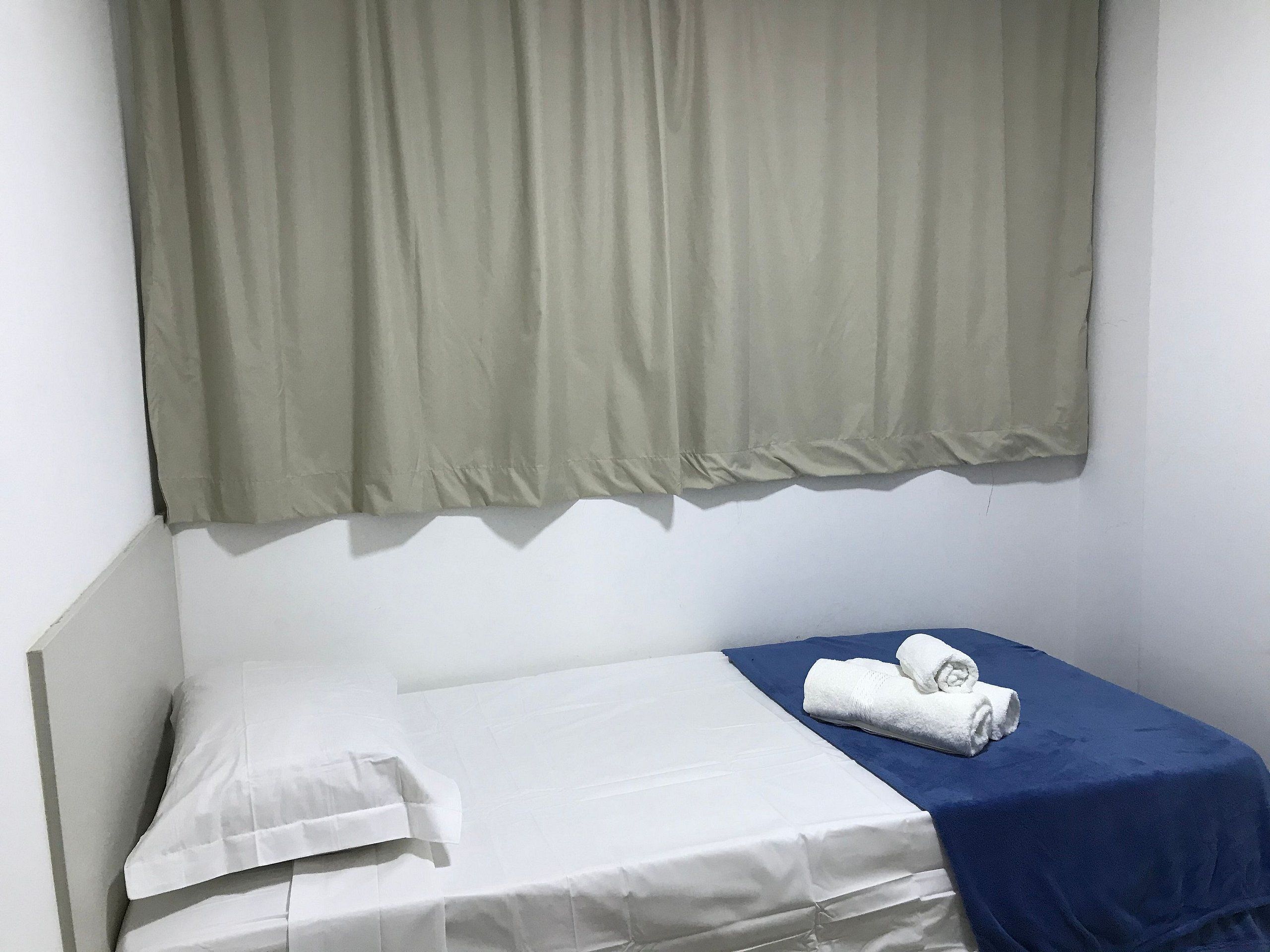 Uma cama de solteiro com roupa de cama branca, um cobertor azul e uma toalha branca enrolada ao lado.