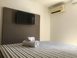 A hotel room with a bed, a flat-screen TV mounted on the wall, and an air conditioning unit.