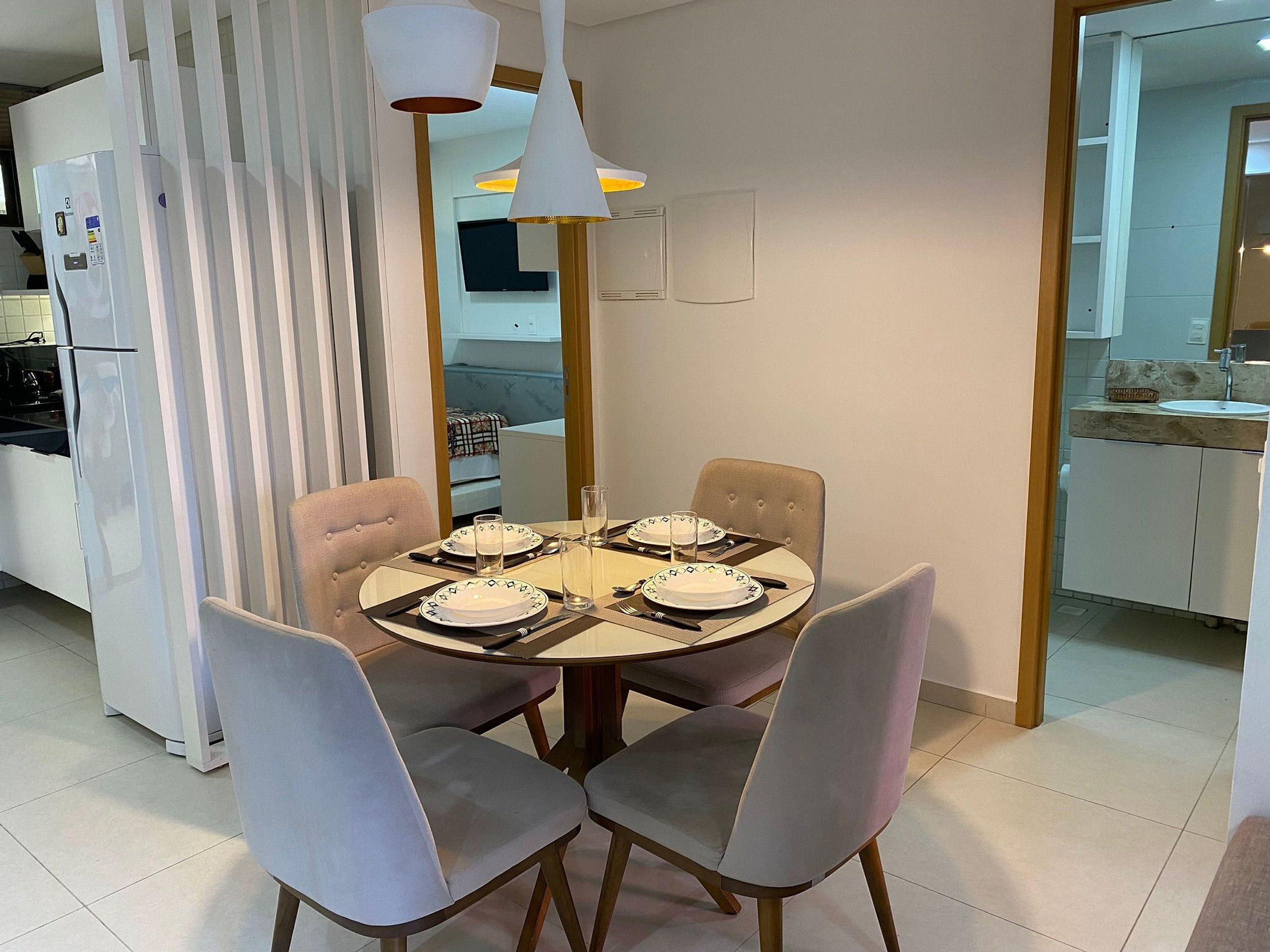 Uma sala de jantar com mesa redonda, quatro cadeiras e cozinha moderna ao fundo.