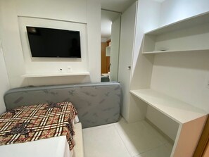 Quarto compacto com uma cama, televisão montada, uma pequena prateleira e uma porta que dá para outro espaço.