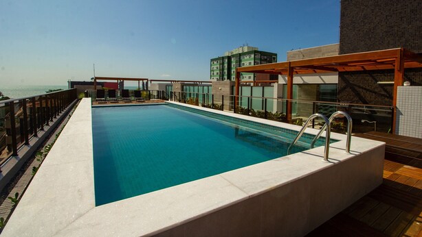 A rooftop swimming pool with a view of the sea, surrounded by modern buildings. - Bela Hospedagem - EcoSummer Flats Tambaú (João Pessoa)