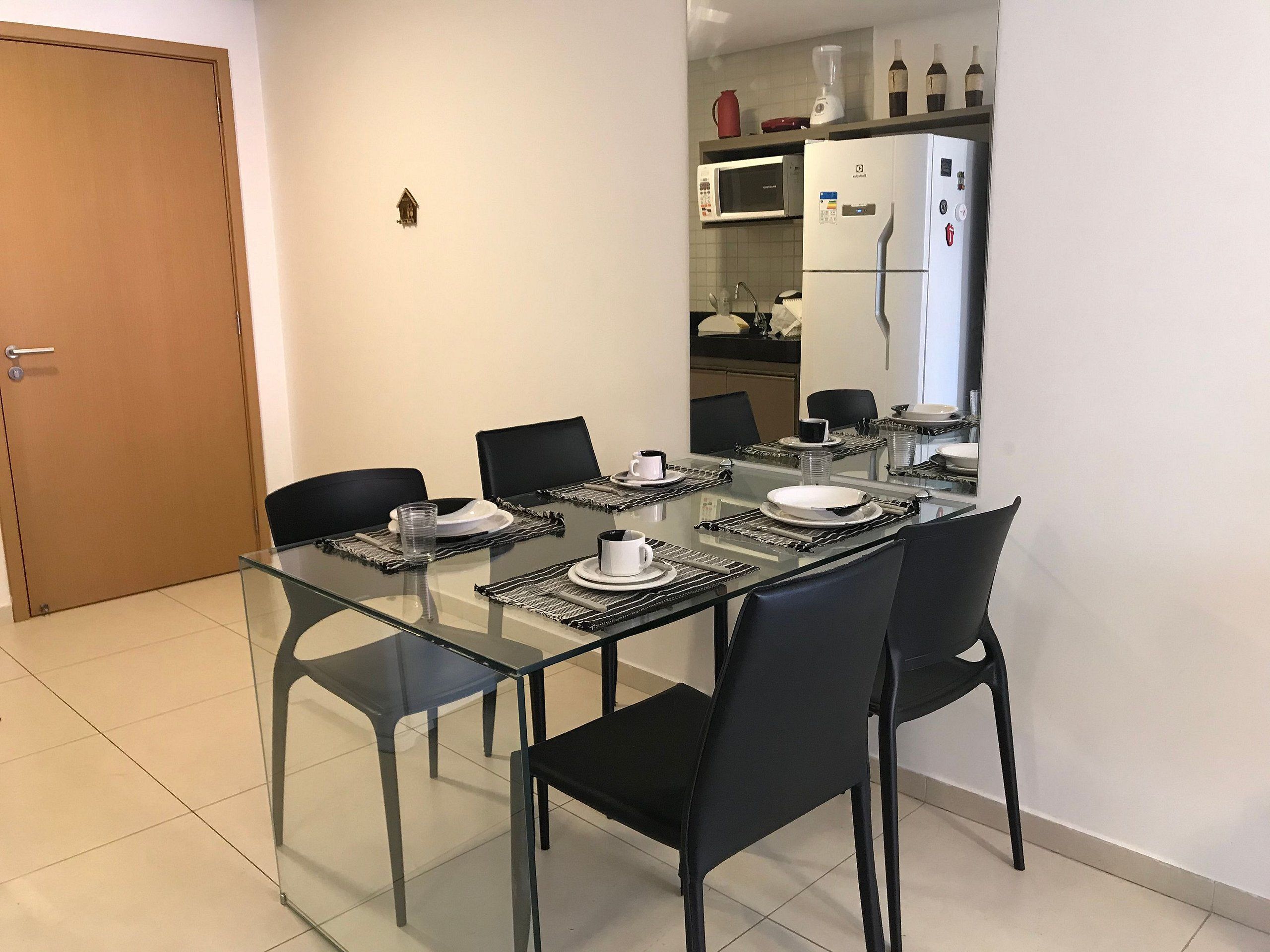 A dining area with a glass table, black chairs, and a kitchen in the background.