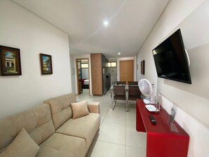 Uma sala de estar com sofá, televisão, ventilador e uma escrivaninha vermelha com um controle remoto.