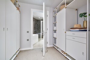 A walk-in closet with white shelving and drawers, a glass shower door, and a plant in a gold vase. - Stay Camden (London)