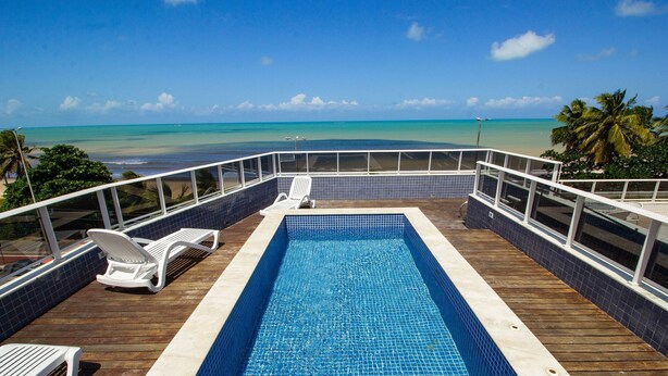 Una piscina con vista al océano, rodeada por una terraza de madera y sillones blancos.