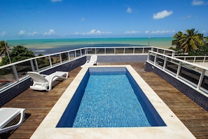 Una piscina con vista al océano, rodeada por una terraza de madera y sillones blancos.