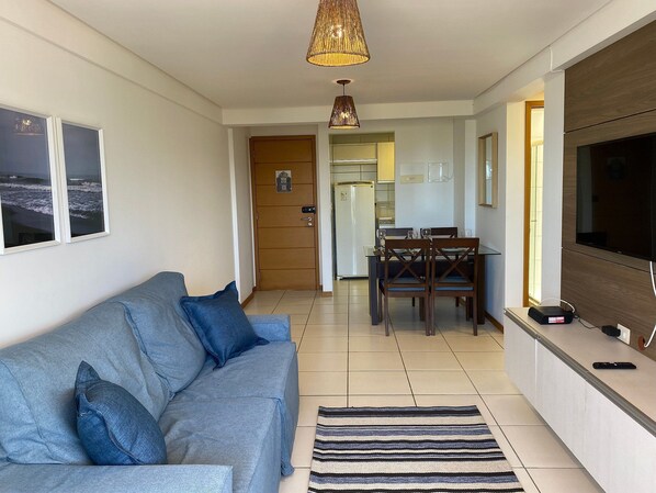 Ein Wohnzimmer mit einer Couch, einem Esstisch mit Stühlen, einem Fernseher und einer Küche im Hintergrund.