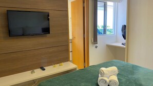 Habitación de hotel con televisión en la pared, cama con manta verde, pared de paneles de madera, puerta y ventana con cortinas.