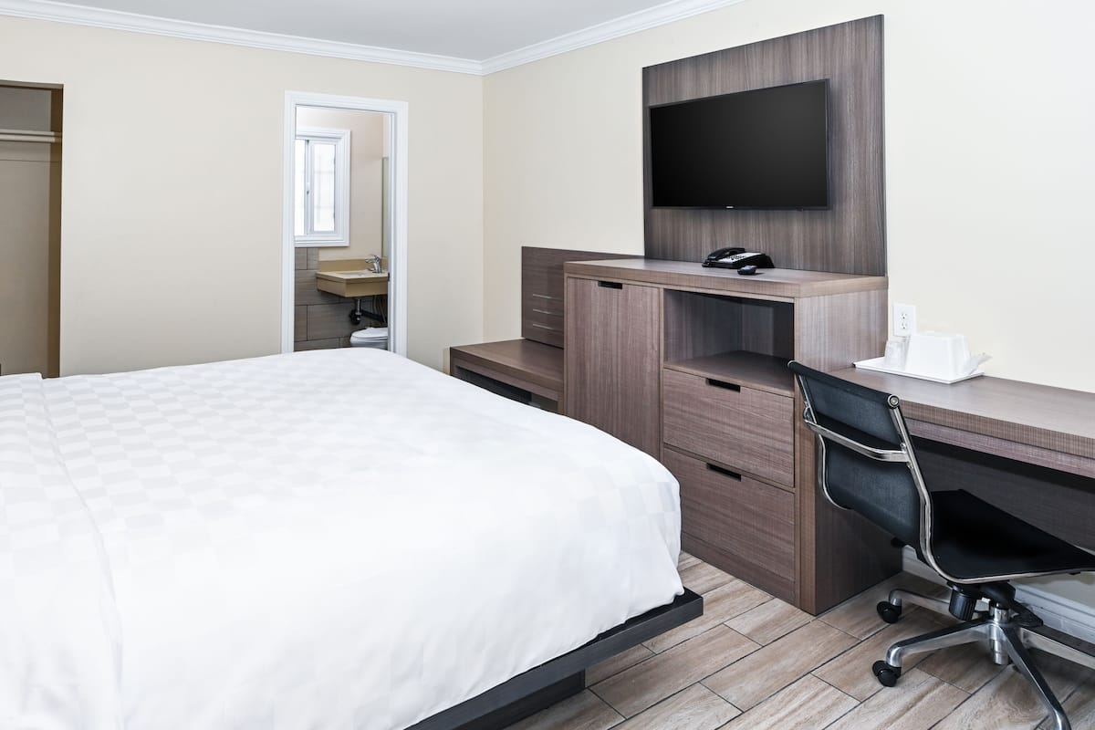 A hotel room with a bed, a desk with a chair, a TV, and a bathroom visible through an open door.