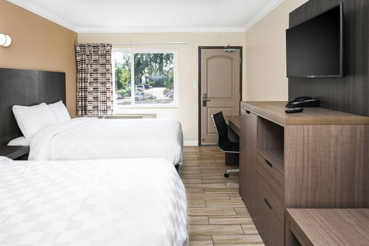 A hotel room with two beds, a desk, a chair, a TV, and a door leading outside.