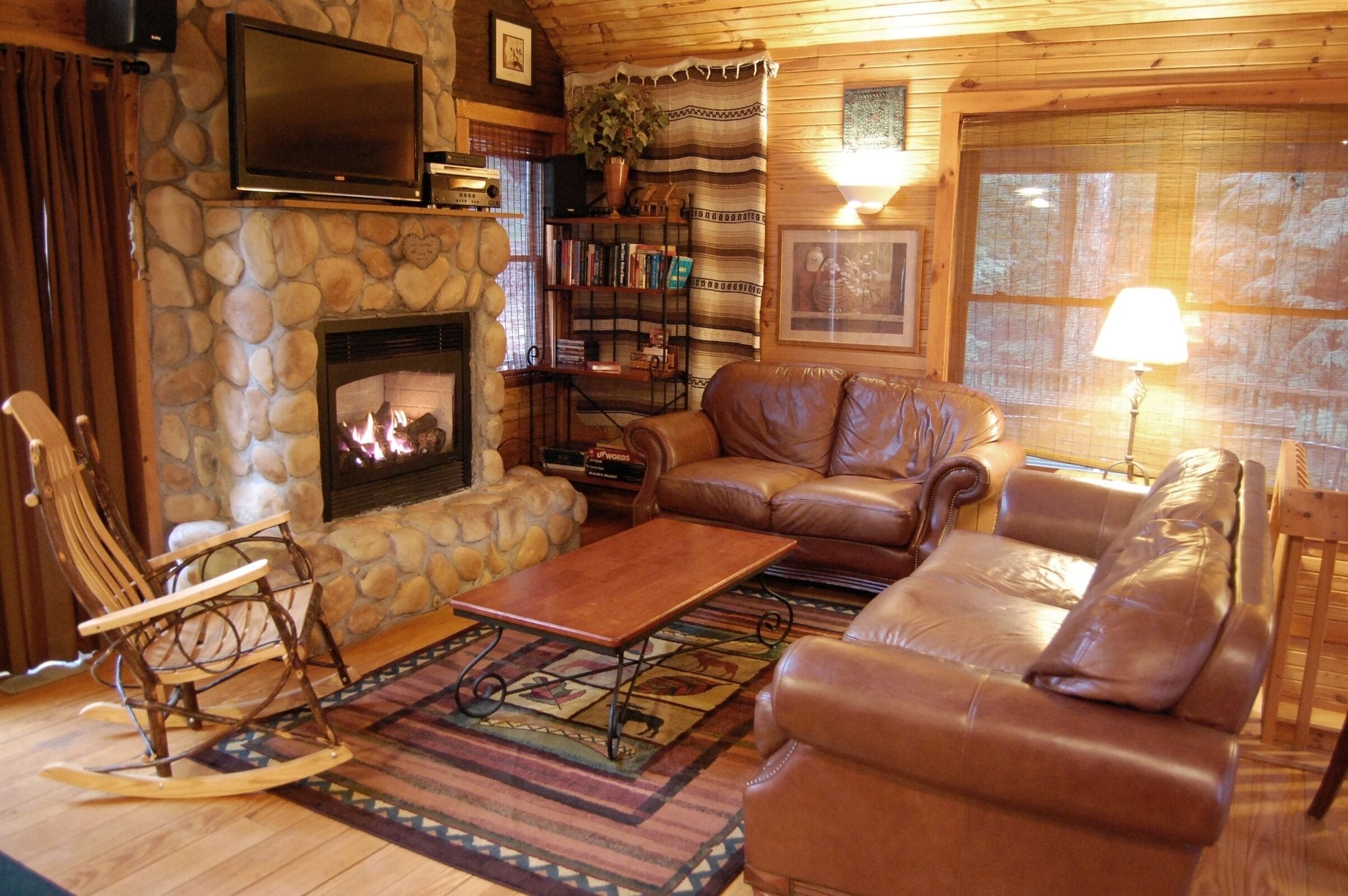 A cozy living room with a stone fireplace, leather furniture, a bookshelf, and a television mounted on the wall.