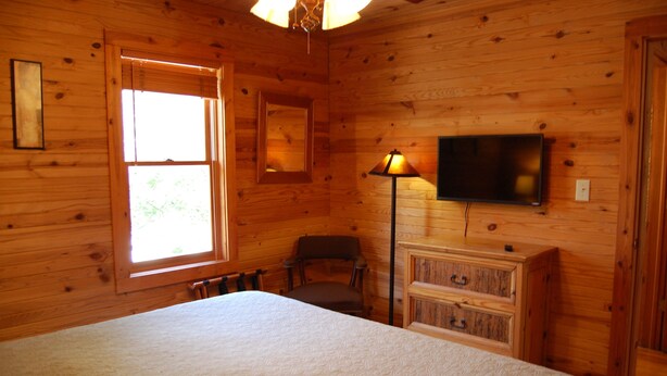 Un dormitorio de cabaña de madera con una cama, una silla, un aparador, un televisor y una ventana.