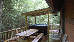 Una terraza de madera con mesas y sillas de picnic, un jacuzzi y una zona cubierta con vistas a un bosque.